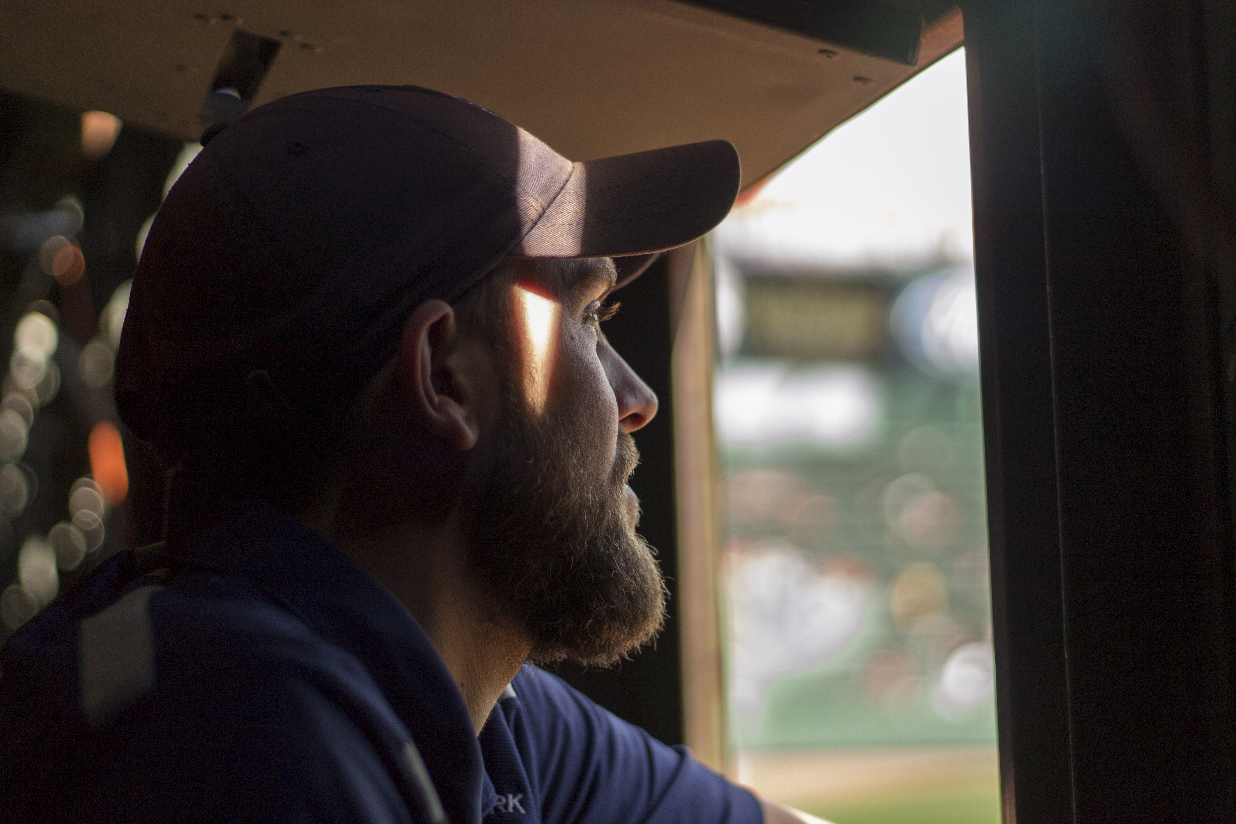 A white man wearing a baseball hat looks out a window onto a baseball diamond.