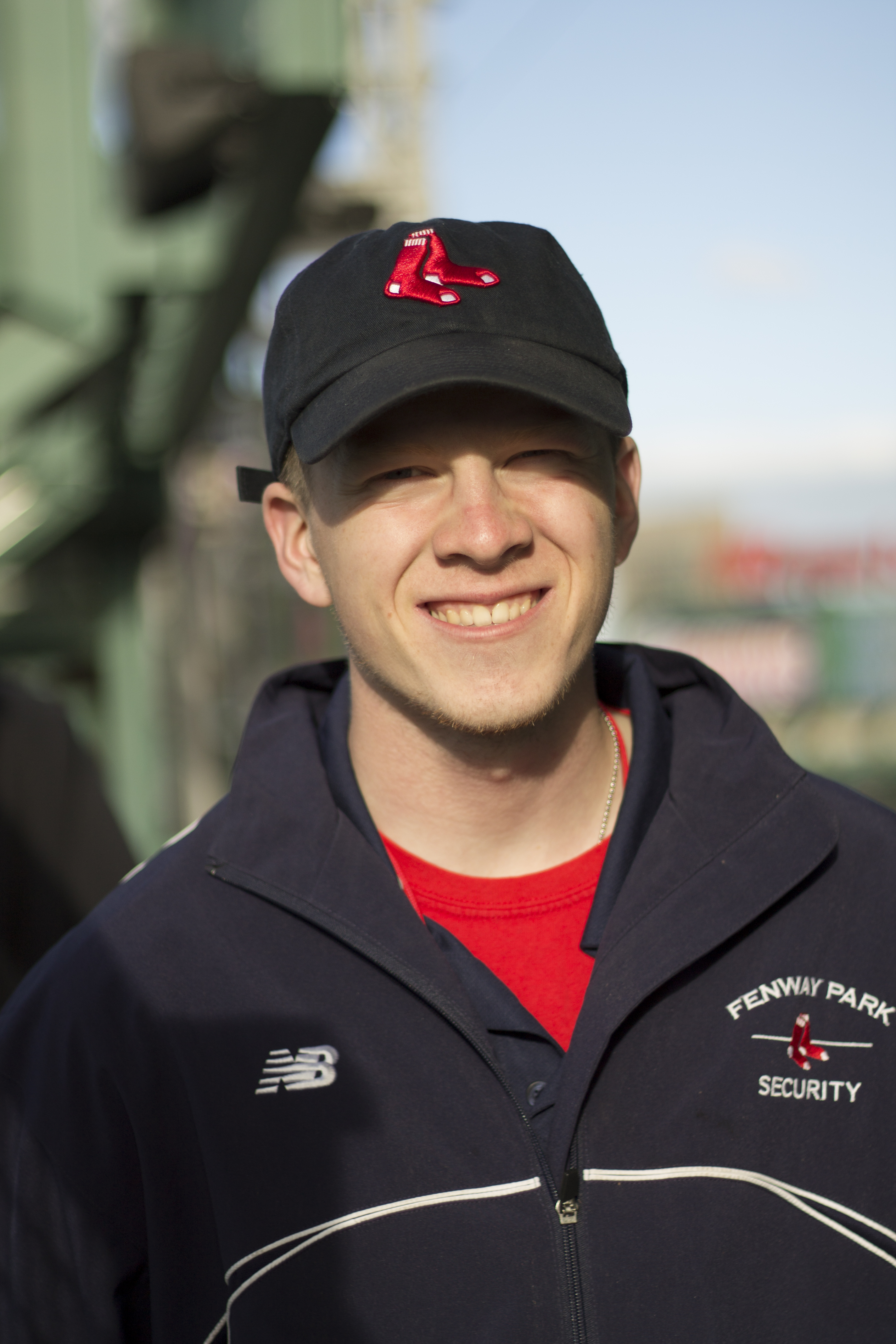 A young white man wearing a navy red sox hat and security jacket smiles at the camera.