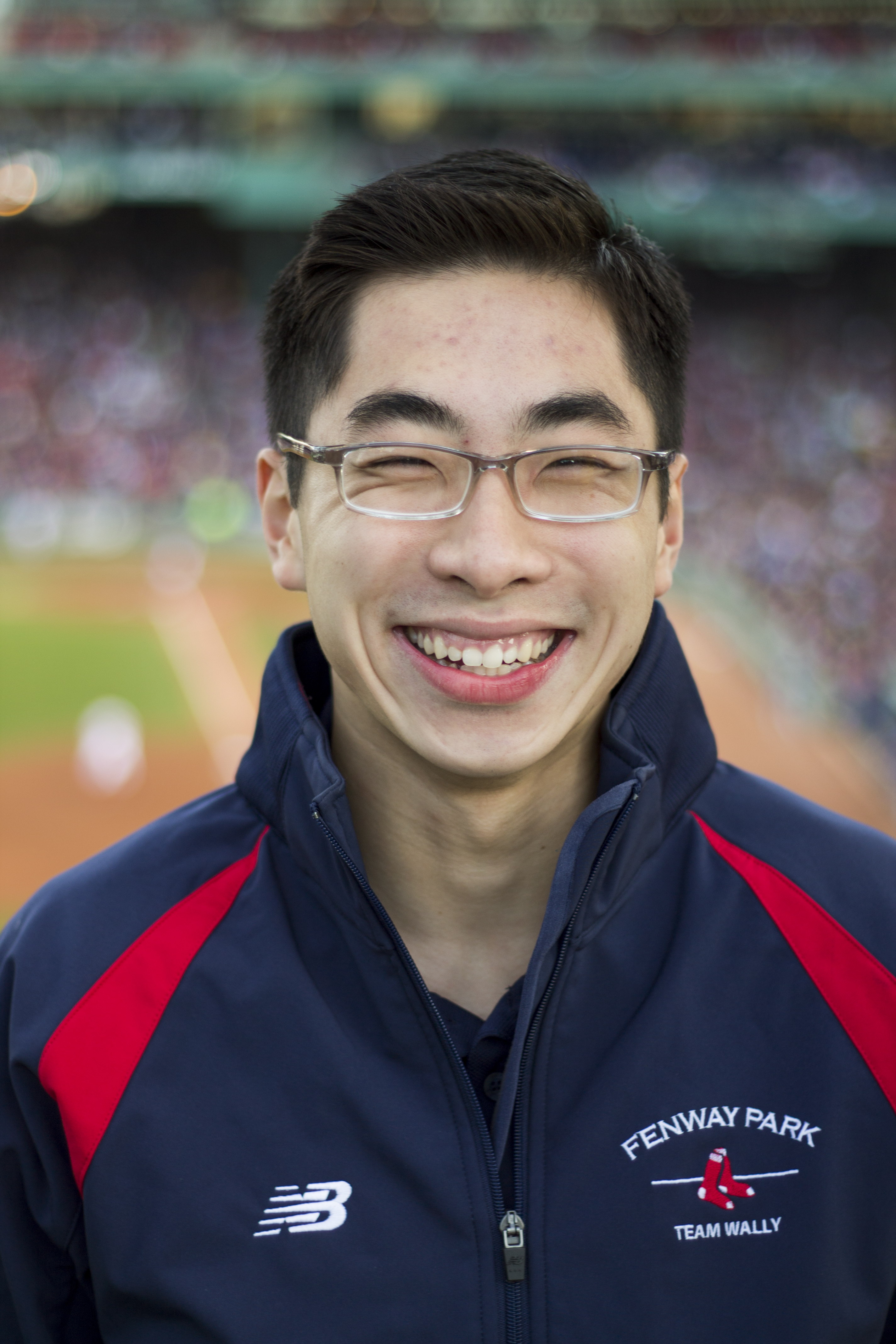 An Asian man wearing glasses and a Red Sox Team Wally uniform smiles at the camera.