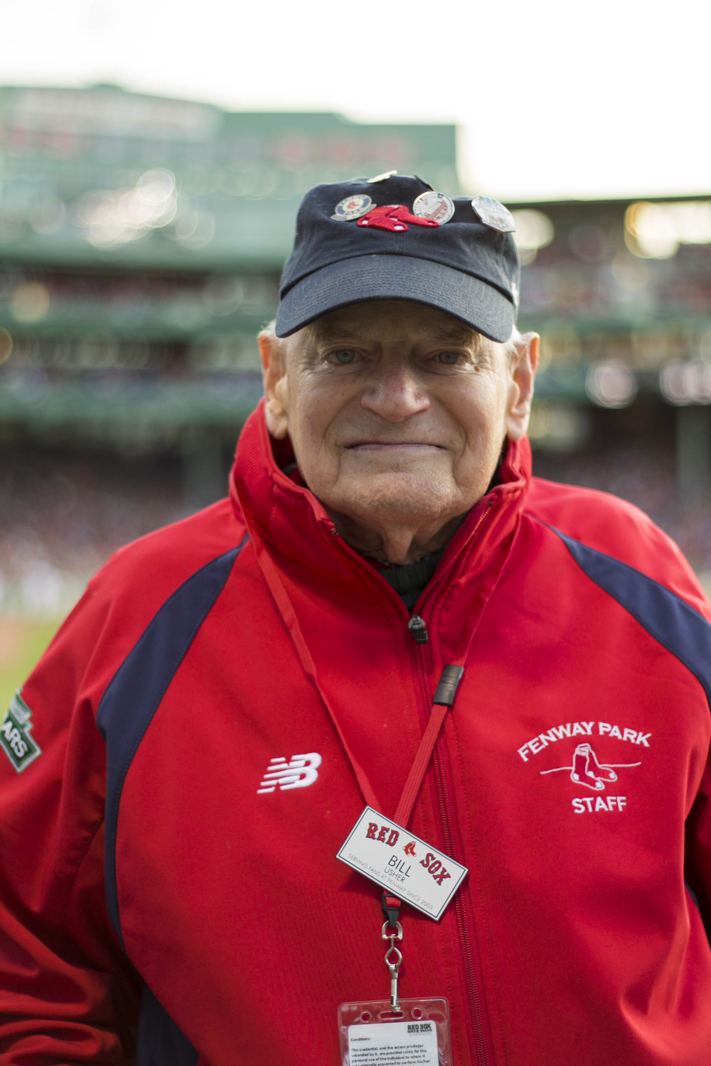An older man wearing a blue hat with pins on it and a red staff jacket.
