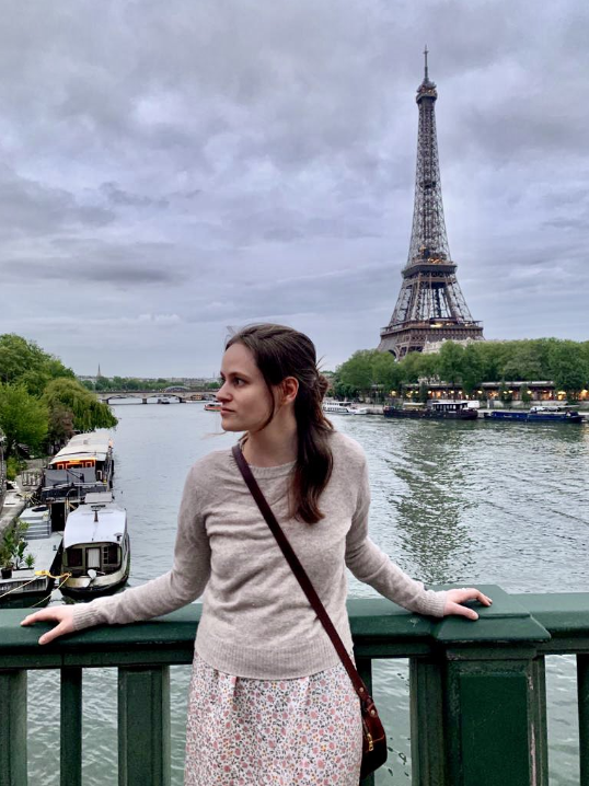 A woman with long brown hair stands in front of the Eiffel Tower.
